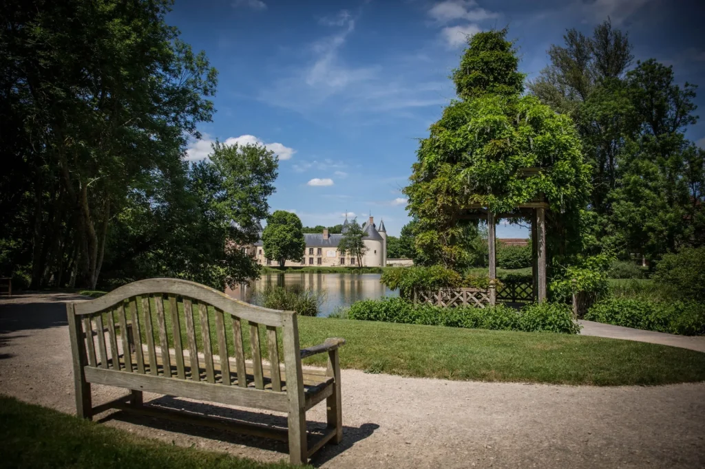 Banc au premier plan dans une allée des jardins avec vue sur le chateau et le plan d'eau